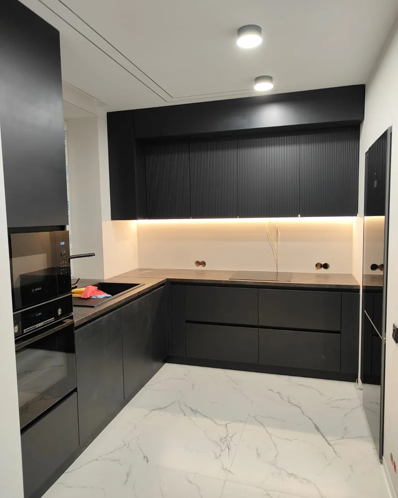 A dark handleless L-shaped fitted kitchen with warm under-cabinet lighting and a pale worktop.