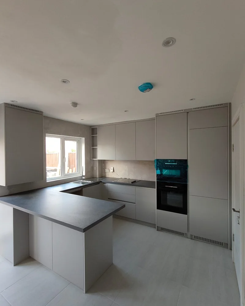 A light grey modern fitted kitchen with a peninsula, integrated oven and dark worktops.