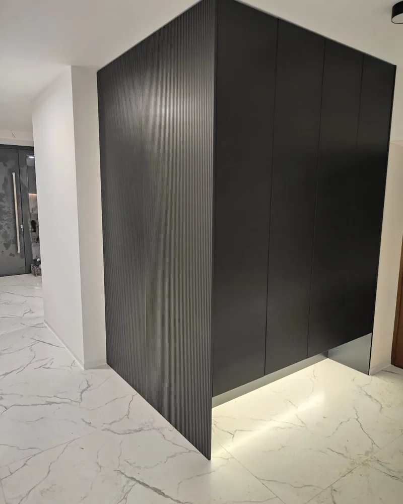 A dark full-height handleless fitted wardrobe with soft plinth lighting in a bright hallway.