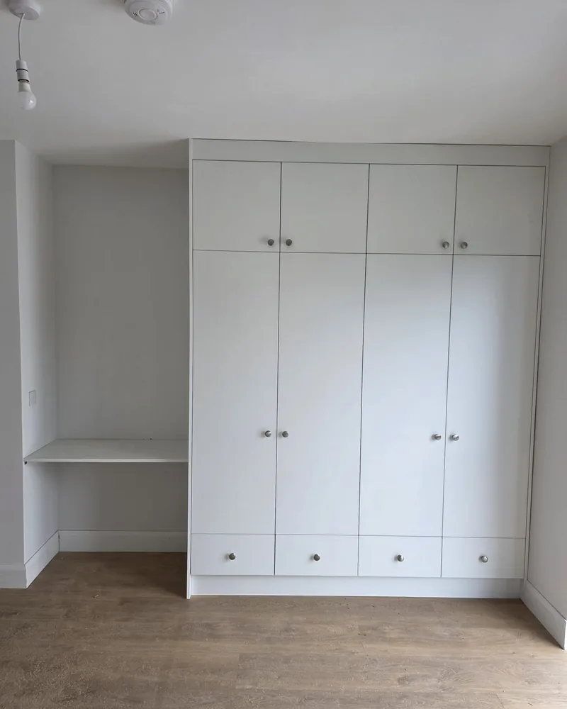 A white fitted bedroom storage wall with wardrobes, overhead cabinets, drawers, shelving and a mirrored sliding section.