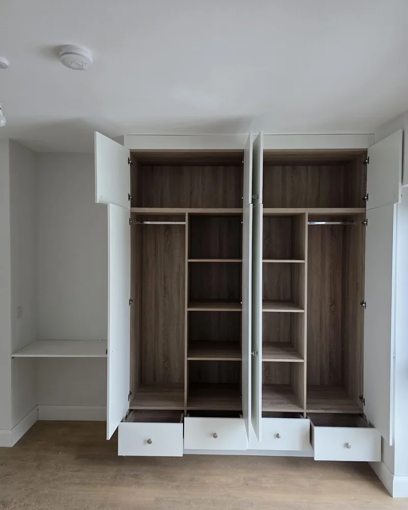 An open white fitted bedroom wardrobe showing internal rails, shelving, drawers and central narrow storage.