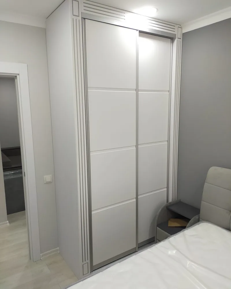 A tall white sliding wardrobe with decorative grooved framing fitted into a compact bedroom.