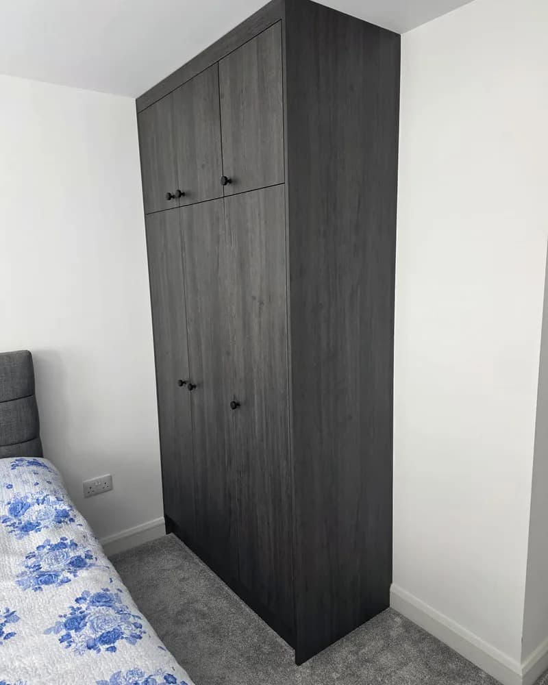 A tall dark wood fitted wardrobe with simple doors and upper storage in a bedroom alcove.