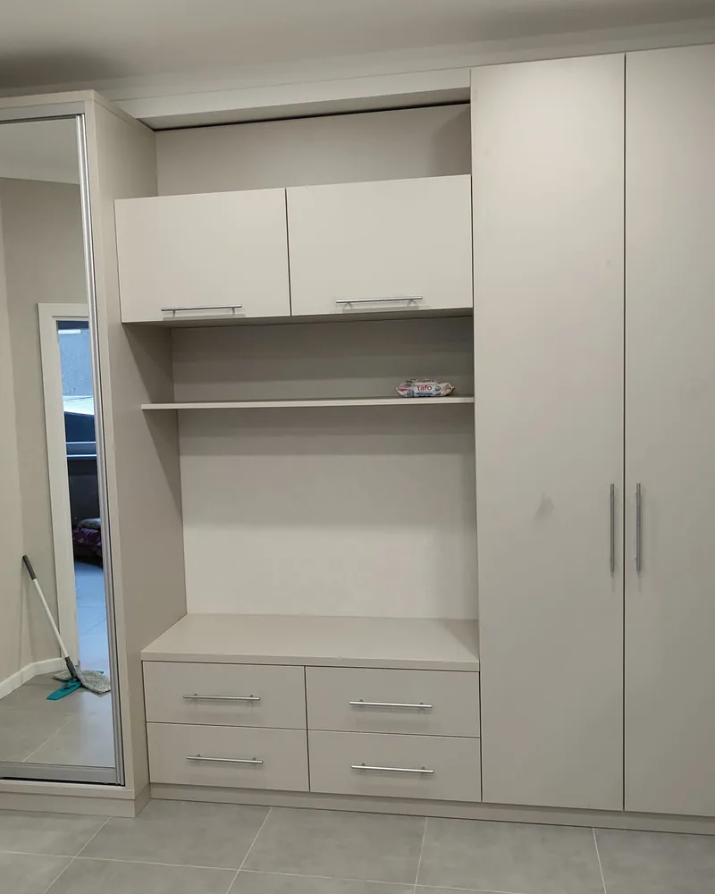 A wide white fitted storage wall with a mirrored sliding wardrobe, overhead cabinets, drawers and open shelf space.