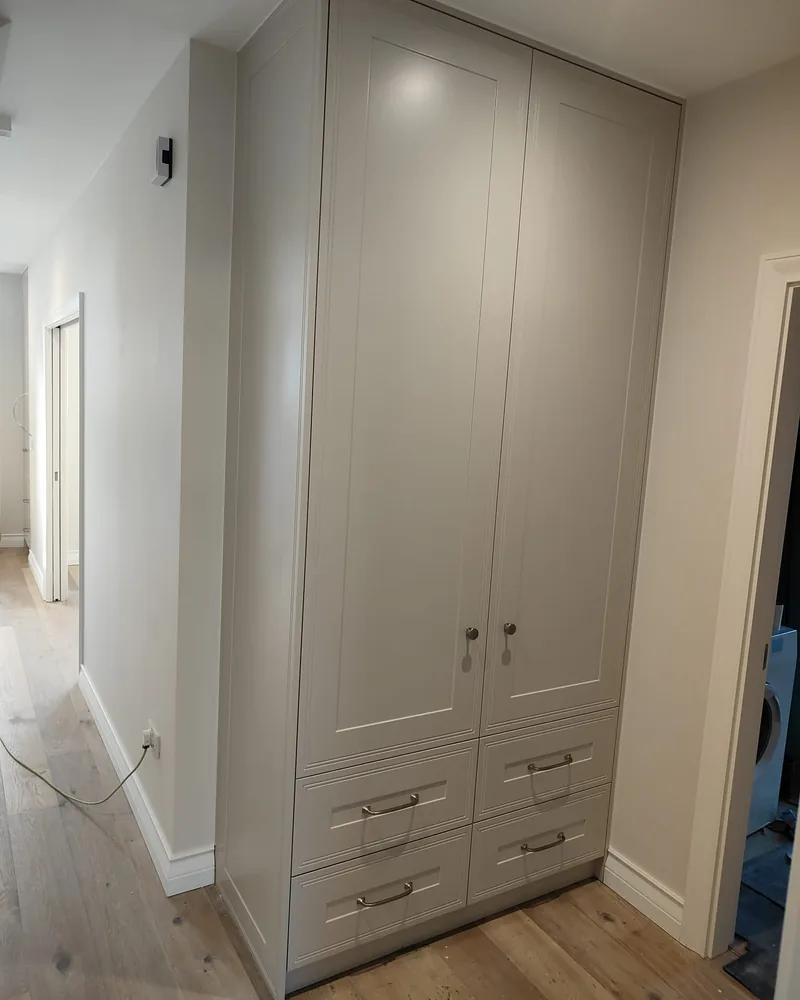 A tall grey fitted corner wardrobe with panel doors, lower drawers and shaped top infill.