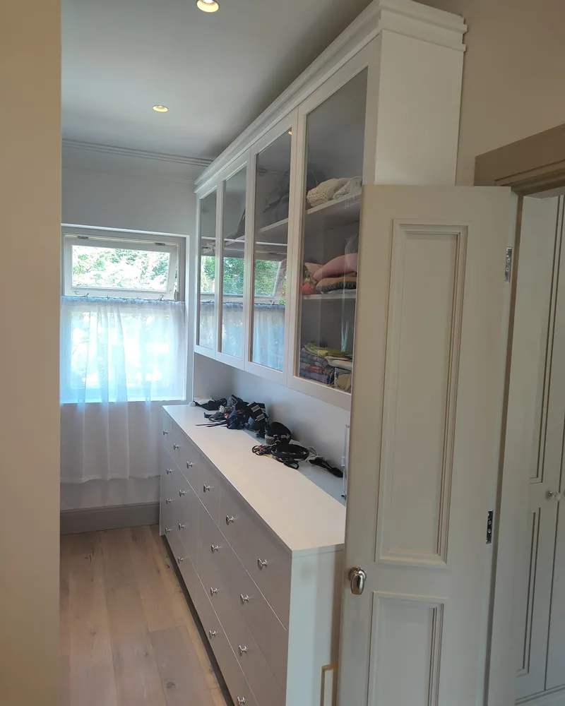 A long white fitted wall combining glazed storage, open shelves, a work surface and tall cabinetry.