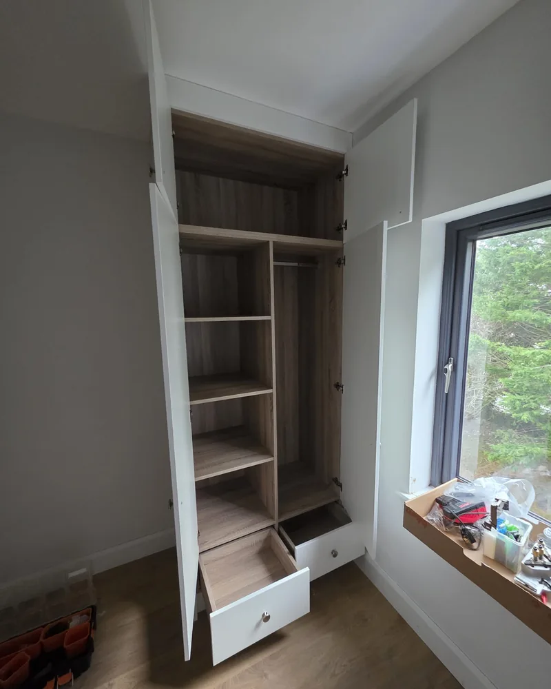 An open window-side fitted wardrobe with shelves, hanging space and lower drawers.